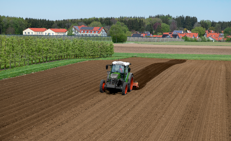 En Fendt e107 Vario arbejder med en kantfræser på en mark ved siden af en frugttræsplantage; huse med solcelleanlæg på tagene kan ses i baggrunden.