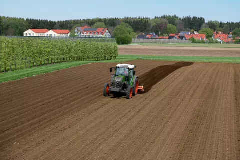 Un Fendt e107 Vario trabaja con un cultivador de caballones en un campo junto a un huerto. Al fondo se ven casas con sistemas fotovoltaicos en sus tejados.