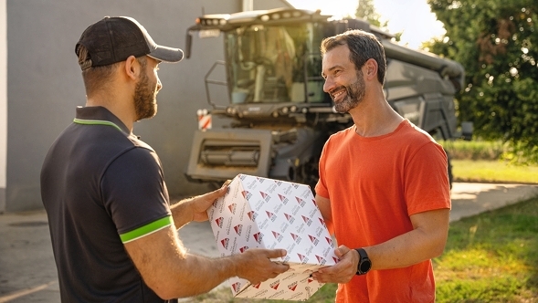 Un trabajador de AGCO Parts entrega un paquete con piezas de recambio AGCO Parts a un granjero para su Fendt IDEAL, que está al fondo.