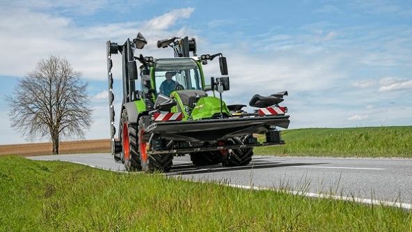 Un tractor Fendt verde circula por una carretera comarcal con la segadora plegada.