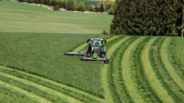 Un tractor Fendt trabajando en un prado verde y siega bajo un cielo lleno de nubes, al amanecer o al anochecer.