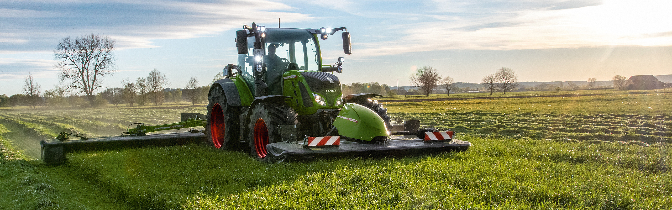 Un tractor Fendt verde trabajando en un prado, rodeado de árboles y bajo un cielo con nubes, al amanecer o al anochecer.