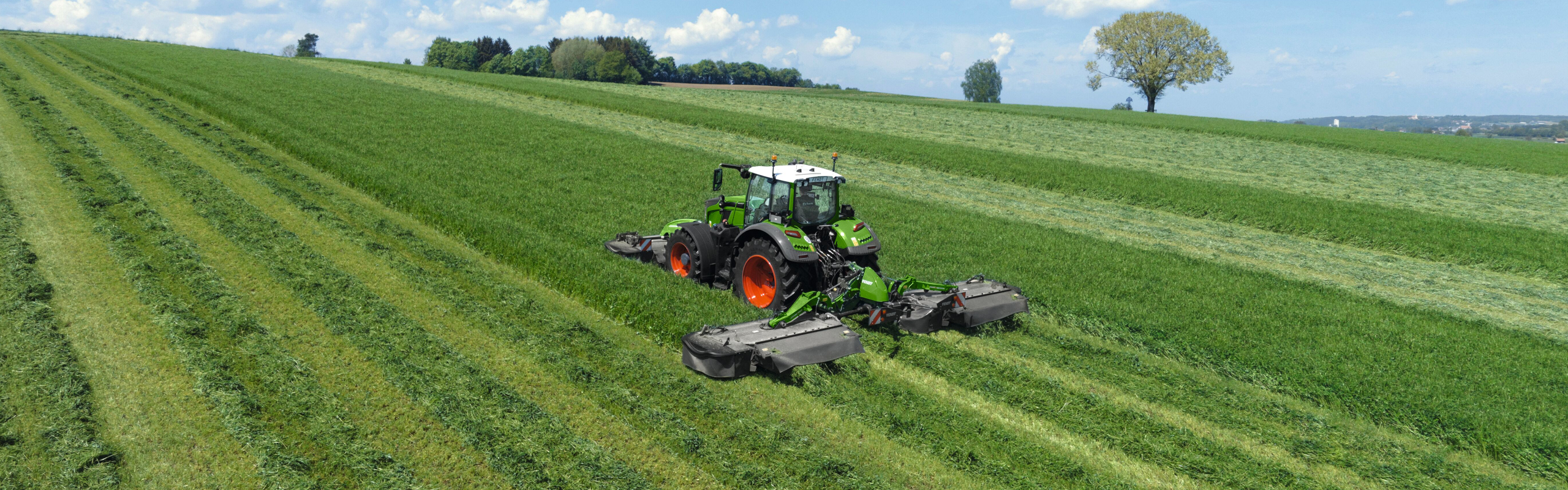 Un agricultor siega con una segadora Fendt Slicer frontal y trasera por prado verde.