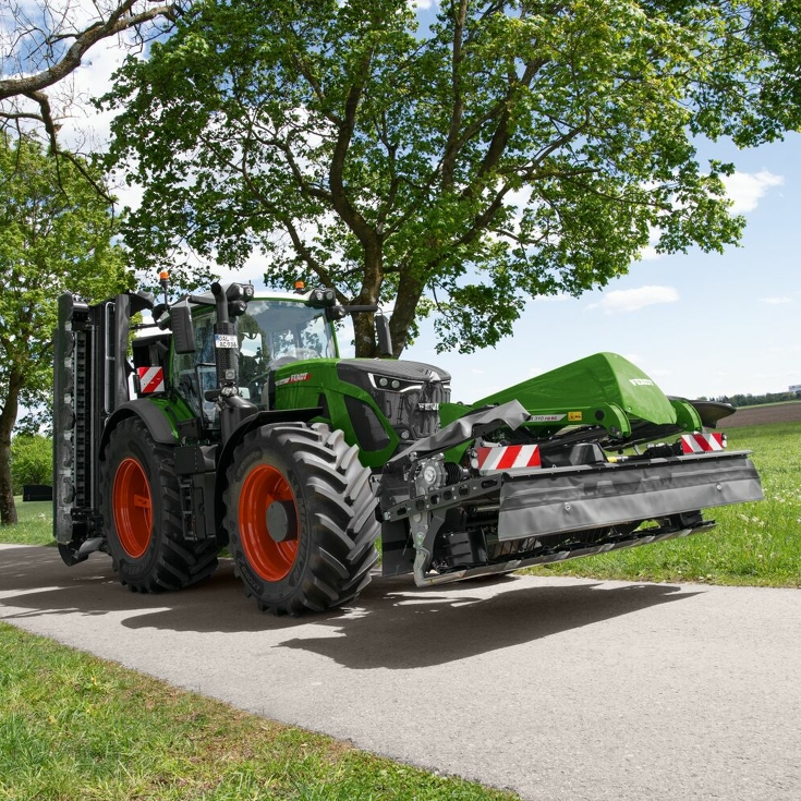 Un granjero conduce por la carretera con un tractor y una segadora frontal y trasera Fendt Slicer, con árboles al fondo.