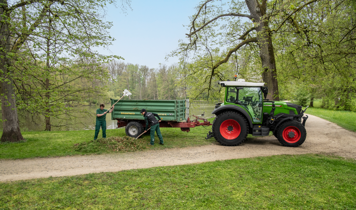 Dos trabajadores de mantenimiento del entorno cargan un remolque detrás de un Fendt e107 Vario con hojas en un parque con un lago rodeado de árboles.