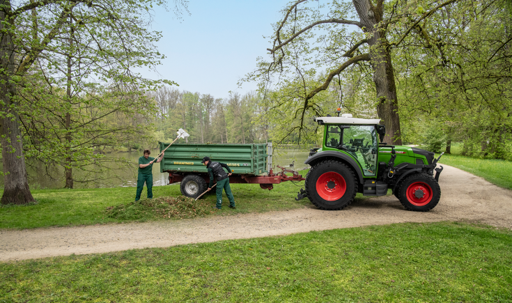 Dos trabajadores de mantenimiento del entorno cargan un remolque detrás de un Fendt e107 Vario con hojas en un parque con un lago rodeado de árboles.