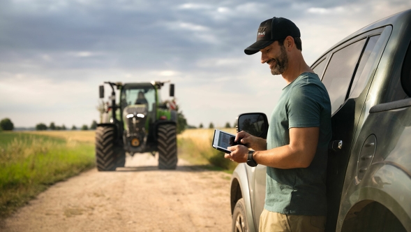 Un agricultor, apoyado en su automóvil al borde del campo, utiliza FendtONE en su teléfono, al fondo se ve un tractor Fendt 700 Vario Gen6.