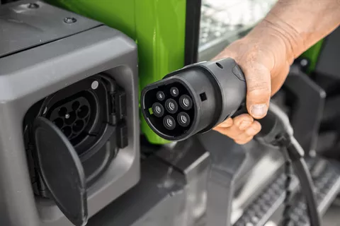 A man's hand holds the plug of the charging cable for the Fendt e100 Vario, next to it is the open charging socket on the tractor