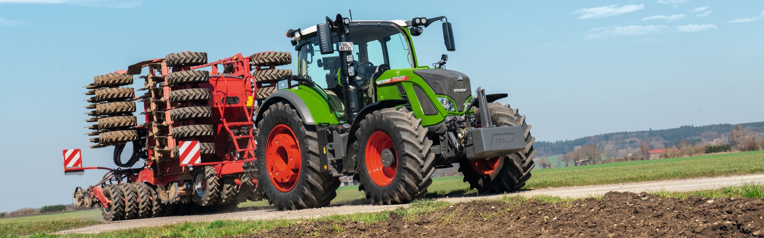 Un agriculteur passe devant un champ sur la route de campagne avec un Fendt 700 Vario Gen7 et un semoir Horsch attelé.