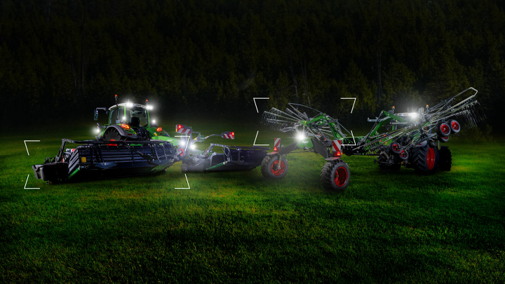 Deux tracteurs équipés d'une faucheuse Fendt Slicer et d'un andaineur Fendt Former sont stationnés dans la prairie au crépuscule et sont éclairés.