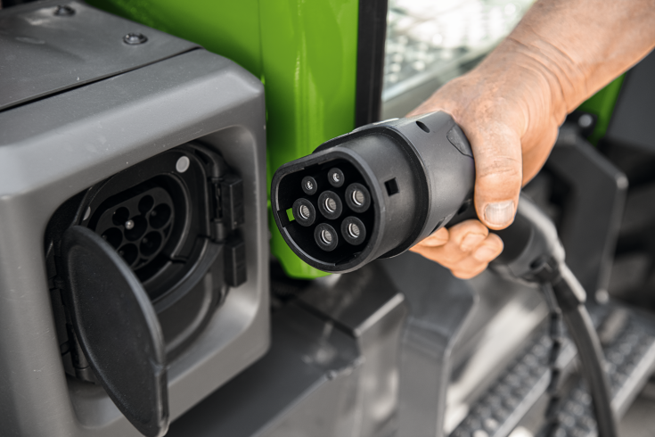 Une main tient la prise du câble de recharge pour le Fendt e100 Vario, à côté de la prise de recharge ouverte sur le tracteur.