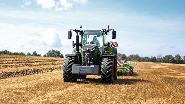 Le Fendt 700 Vario Gen 7 en action dans un champ. En arrière-plan, des arbres et un ciel bleu avec des nuages blancs.