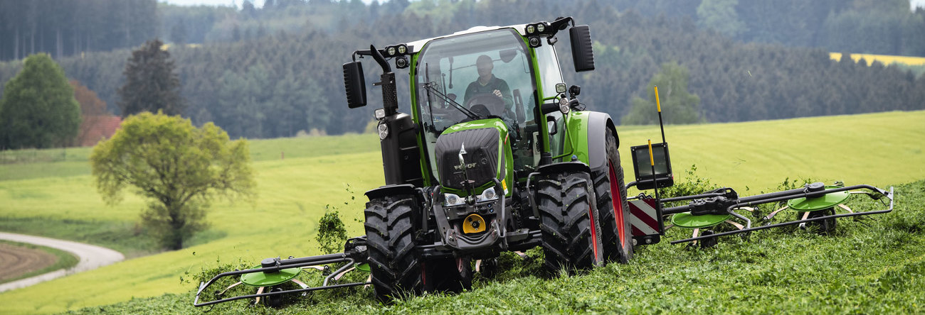 egy zöld Fendt traktor piros felnikkel és a zöld Fendt Lotus munkaeszközzel egy zöld mezőn a fű betakarítása közben, a tájra nyíló panorámával.