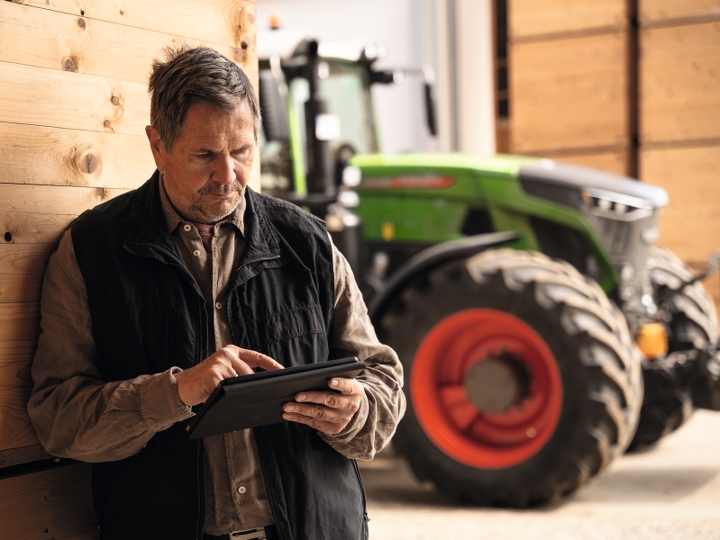 Egy gazda faborítású fal előtt áll, traktorja mellett, kezében egy tablettel