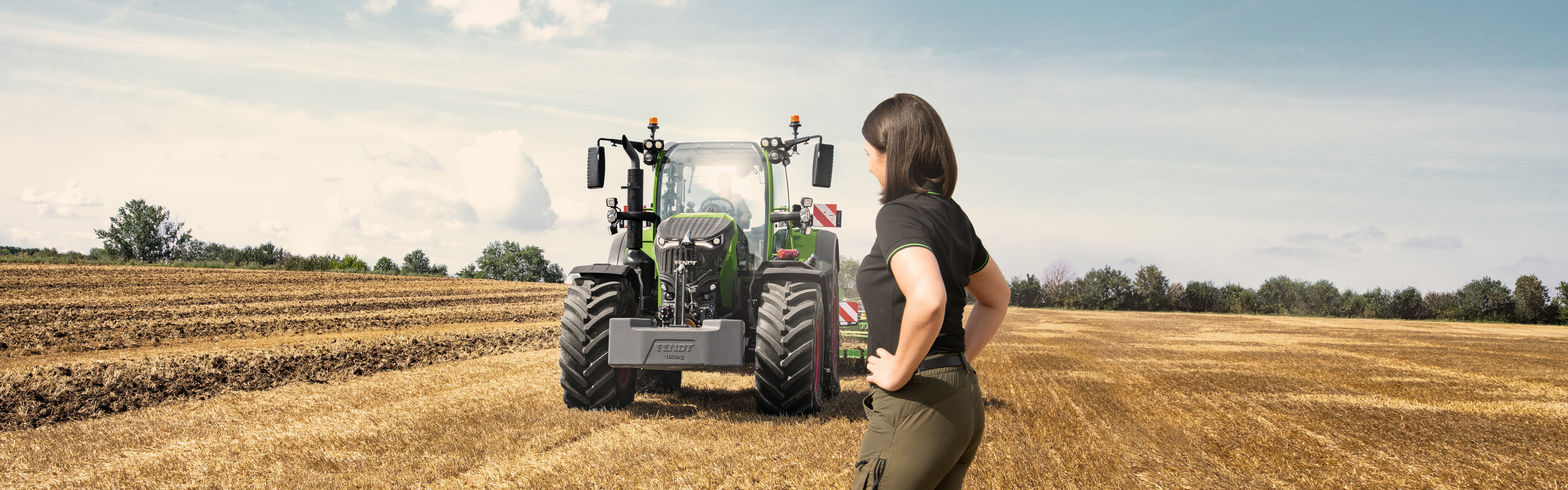 Egy gazdaasszony a szántóföldön áll az új Fend 700 Vario Gen7 traktorja előtt.