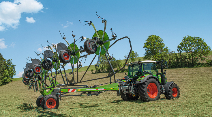egy zöld Fendt traktor piros felnikkel és a zöld Fendt Lotus-szal összecsukott állapotban egy zöld mezőn kék égbolttal és kilátással egy zöld dombra.