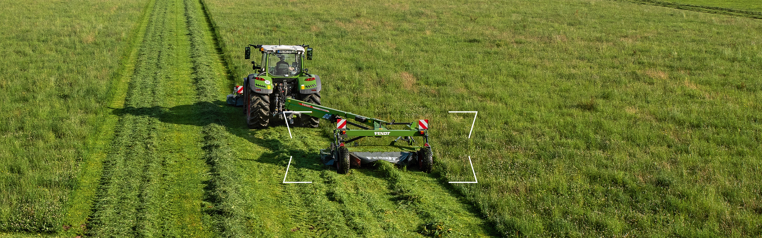 Vontatott Fendt Slicer transzportfutóművel egy nagy mező kaszálása közben. A háttérben erdő látható.