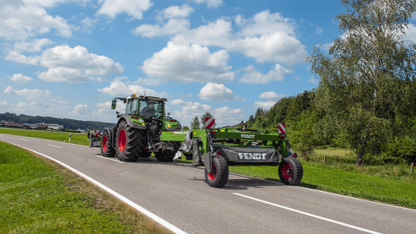 Egy zöld Fendt traktor egy vontatott Fendt Slicer transzportfutóműves kaszával az országúton. A háttérben kék ég, fehér felhők és egy erdő látható.