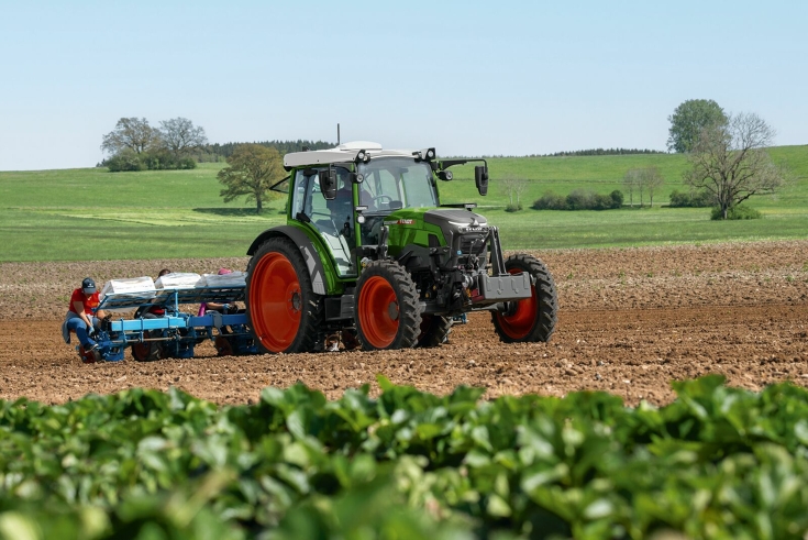 A Fendt e100 Vario is driving over a field, with a planting machine and planters in the rear for planting work, in the foreground strawberry plants