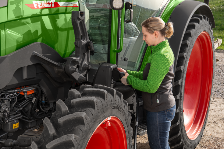 A young woman with a Fendt jacket plugs the Fendt e100 Vario into the charging cable to charge it.