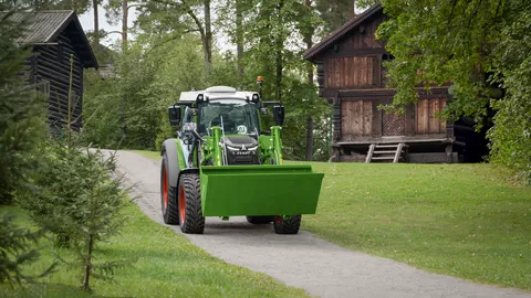 A Fendt e107 Vario driving down a road in Norway with a Fendt frontloader and bucket, wooden houses in the background