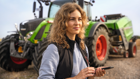 A farmer in front of her Fendt agricultural machinery opening the Fendt News app on her mobile phone