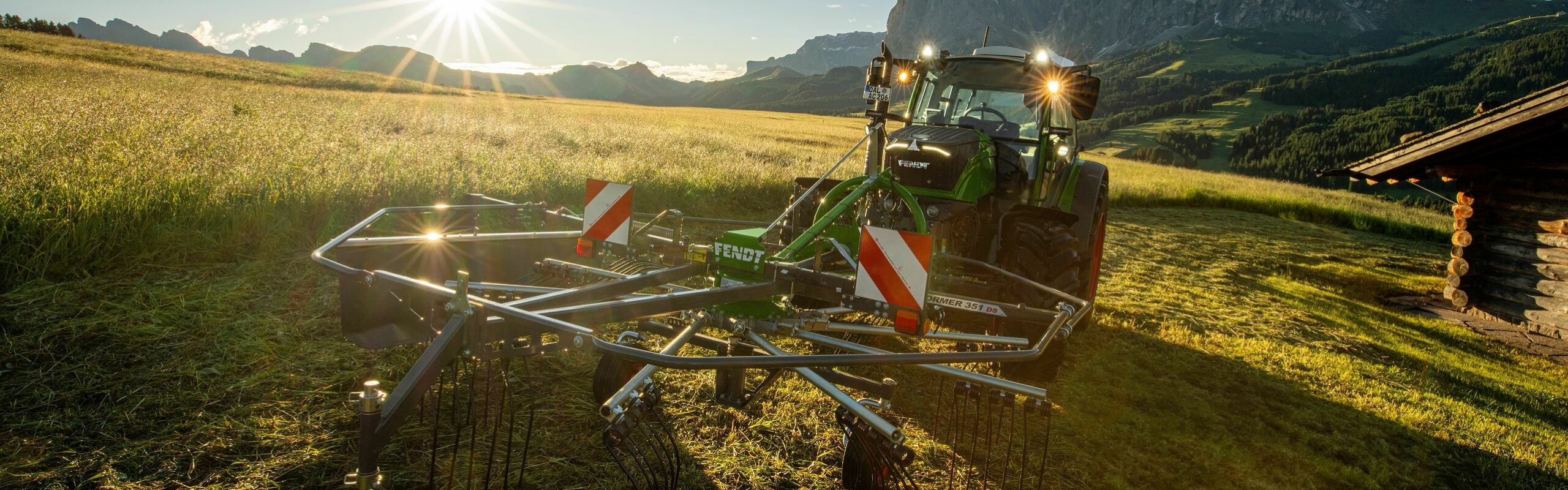 The green Fendt tractor with red rims and the green Fendt Former Alpine implement in use on a mountain meadow. In the background is a mountain landscape, a blue sky and the radiant sun.