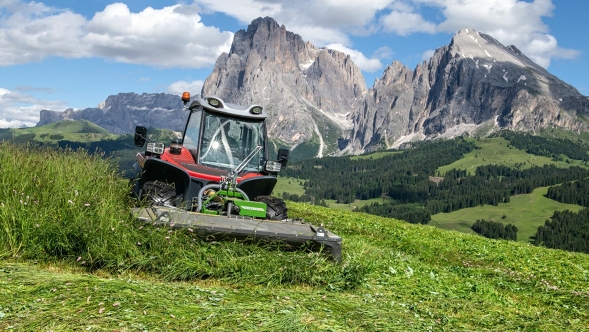 A tractor with the Fendt Slicer Alpine front mower drives on steep terrain and mows a mountain meadow. In the background is a blue sky with white clouds and large mountains.