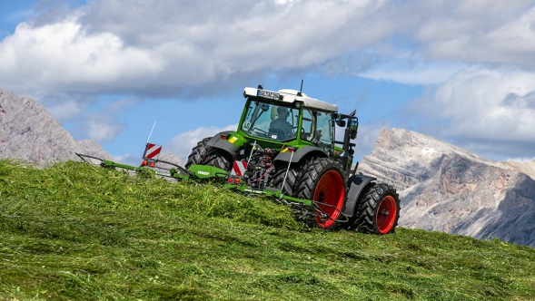 The green Fendt tractor with red rims and the Fendt Twister Alpine tedding the forage in a mountain meadow.  In the background is a blue sky with white clouds and large mountains.