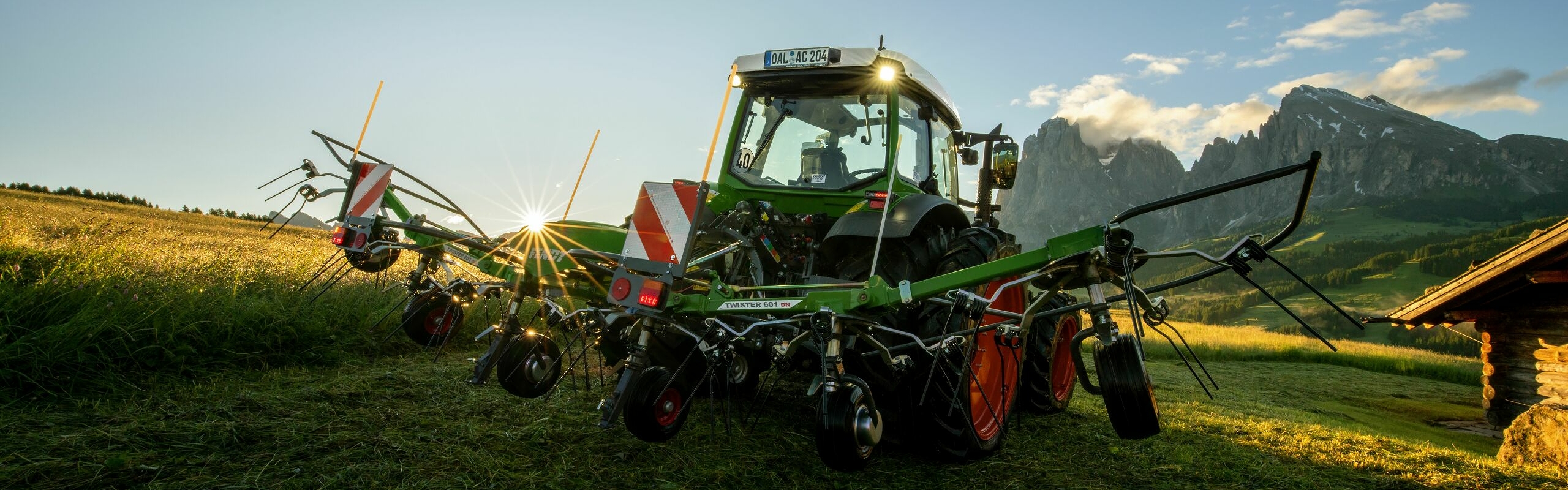 The green Fendt Twister 601 DN with a green Fendt tractor and red rims standing on a mountain meadow. In the background are high mountains and the sun is rising