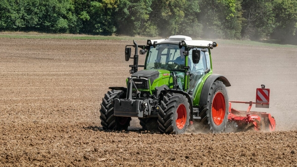 Fendt 200 Vario working in the field. Dust swirls and green trees can be seen in the background.
