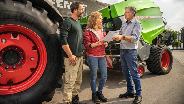 A farmer and a farmer's wife stand in front of a tractor with a round baler and receive advice from a dealer.