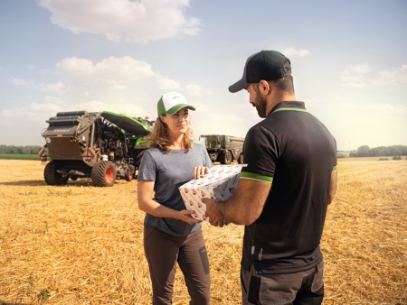 A farmer stands in front of a crop field and accepts a package of AGCO Parts handed to her by a Fendt service employee.