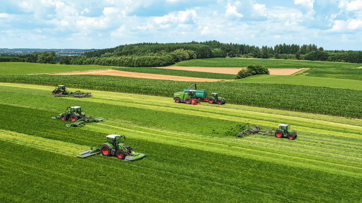 6 Fendt tractors and a Fendt Katana forage harvester are staggered on a green field, mowing, turning, raking and choppin
