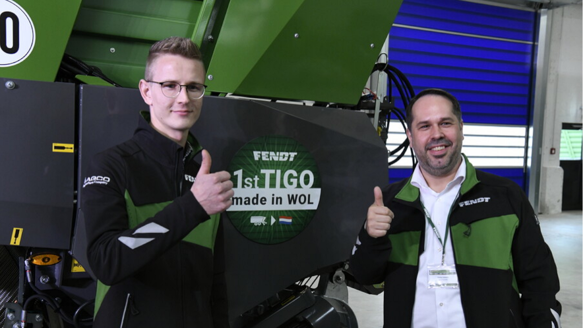 Two Fendt employees stand in front of a Fendt Tigo and hold up their right thumbs. A sticker with the text ‘1st Tigo made in WOL’ is stuck on the Tigo.