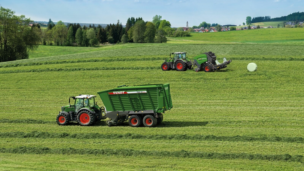 Two farmers are each driving a tractor in a meadow, one is baling round bales with a Fendt Rotana, the other is collecting hay with a Fendt tractor.