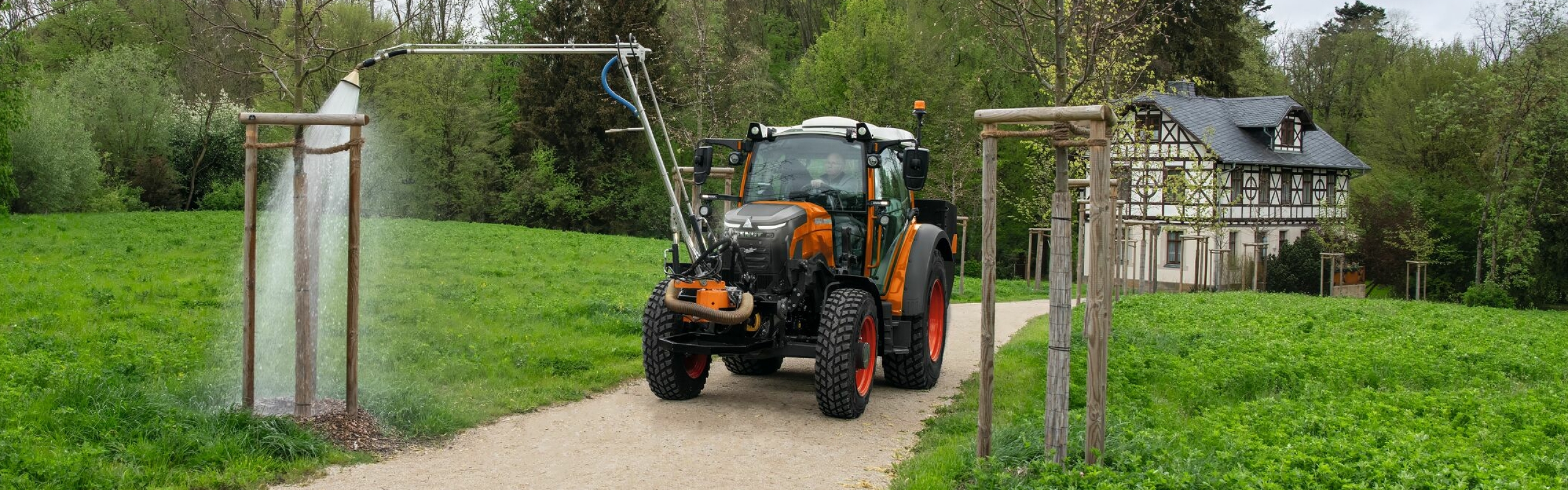 An employee driving an orange-painted ISE Fendt e100 Vario tractor and watering trees in a community