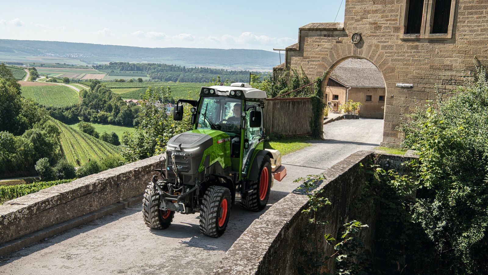 A green Fendt e100 V Vario driving out of a yard above the vineyards over a bridge