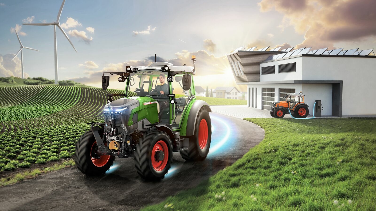 A Fendt e100 Vario travelling along a country lane near the town, with wind turbines running in the background and another Fendt e100 charging