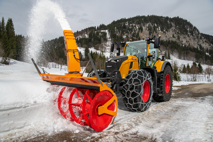 A Fendt 728 Vario with snow chains is clearing snow from a road, with a mountain of conifers in the background