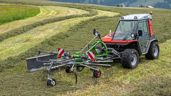 The green Fendt Former Alpine rake is in use on a mountain meadow harvesting forage.  In the background you can see the rake tracks on the meadow.