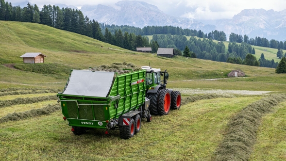 A green Fendt tractor with red fields driving on a meadow with the green Fendt Tigo ST forage wagon. In the background are hills, trees and mountains under a cloudy sky.