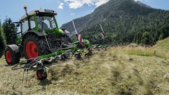 The green Fendt Twister with a green Fendt tractor and red rims tedding hay on a mountain meadow with a blue sky and mountain in the background.