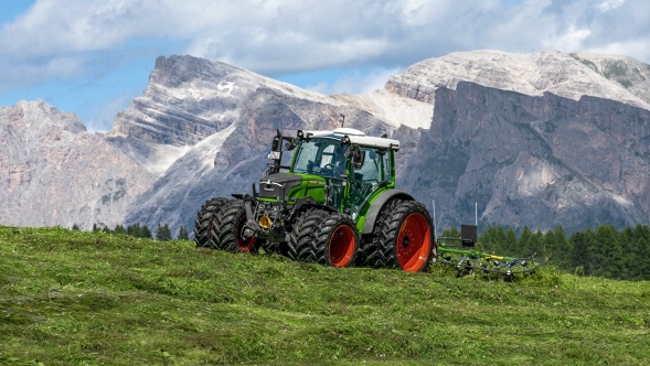 A green painted Fendt 200 Vario with attached Fendt Alpine tedder in alpine use in front of a massive mountain backdrop.