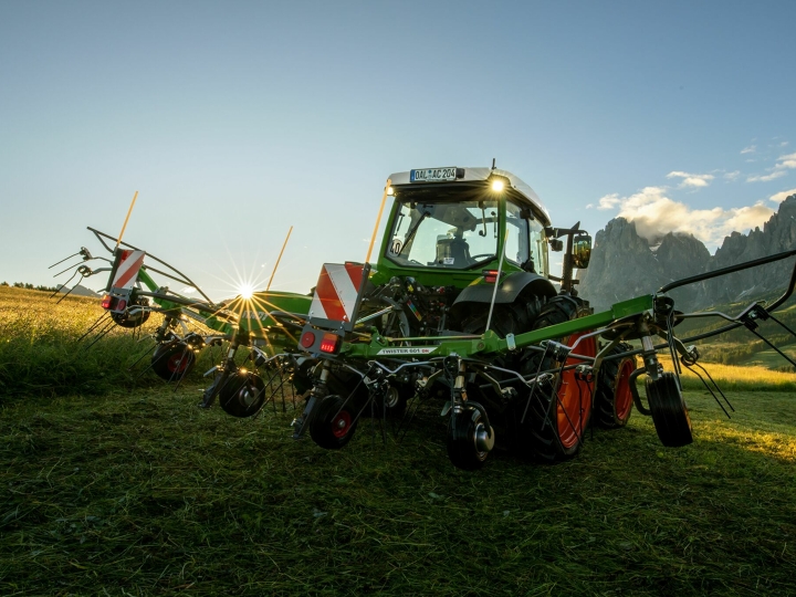 The green Fendt Twister hay tedder standing in front of a green Fendt tractor in a mountain meadow. In the background, the sun is slowly setting, the sky is blue and the mountains can also be seen