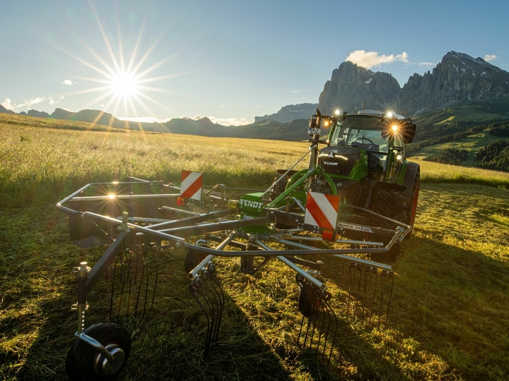 The green Fendt Former Alpine rake stands in front of a green Fendt tractor in a mountain meadow. In the background, the sun is slowly setting, the sky is blue and the mountains can also be seen