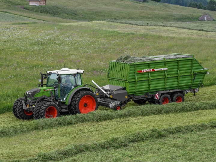 A green Fendt tractor with red rims and a green Fendt Tigo forage wagon with red rims driving on a green mountain meadow