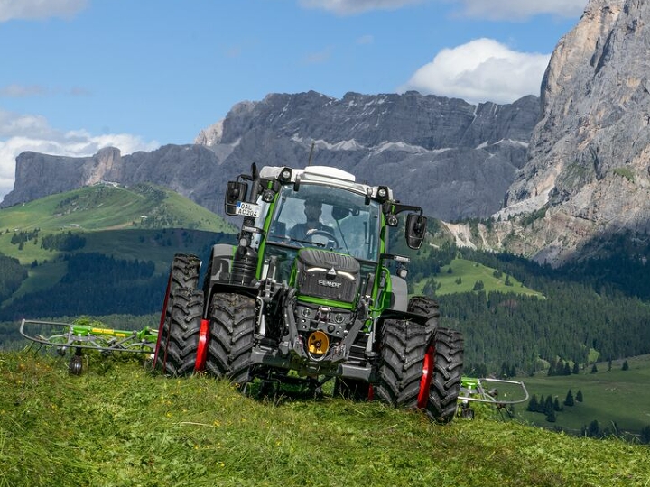 The Fendt 200 Vario with double tyres and red rims driving on a green mountain meadow, with mountains and blue sky in the background