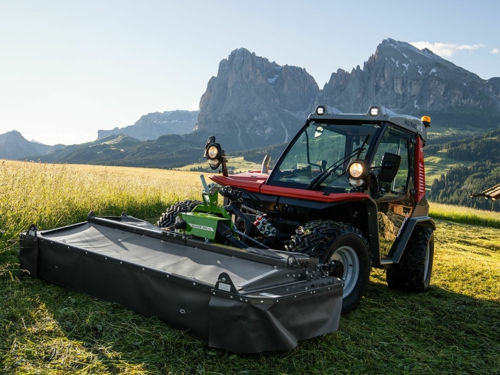 The Fendt Slicer Alpine mower in action on a mountain meadow with mountains and blue sky in the background