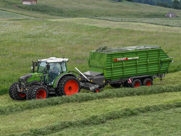 A green Fendt tractor with red rims and a green Fendt Tigo forage wagon with red rims driving on a green mountain meadow